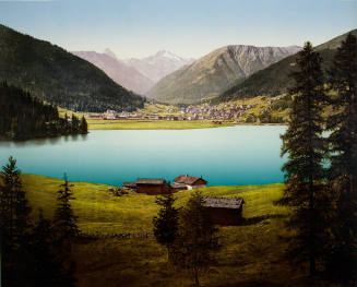 Mountain landscape with a turquoise lake, green meadows, cabins, a village, and snow-capped mou…
