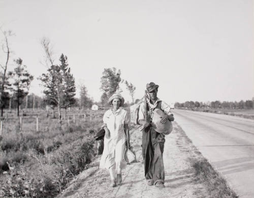 A man and woman walking along the highway. They say "Damned if we'll work for what they pay folks hereabouts."