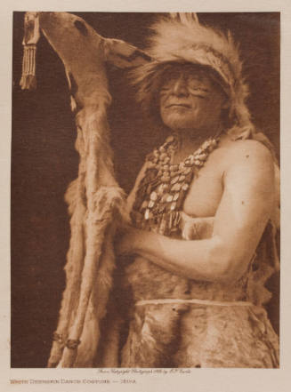 Person in traditional Native American attire with feather headdress, shell necklaces, and holdi…